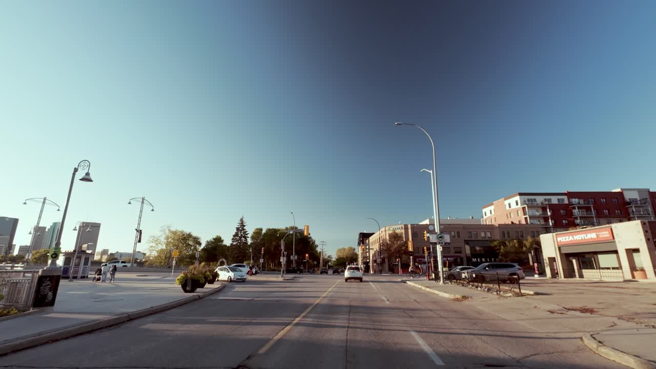 POV shot from car driving down empty street; river left, trees right; city skyline ahead; fall season