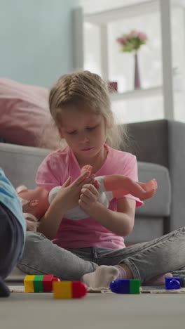 toddler hits constructor with airplane while preschooler girl rocking baby doll in arms. siblings play with toys sitting on floor at home closeup