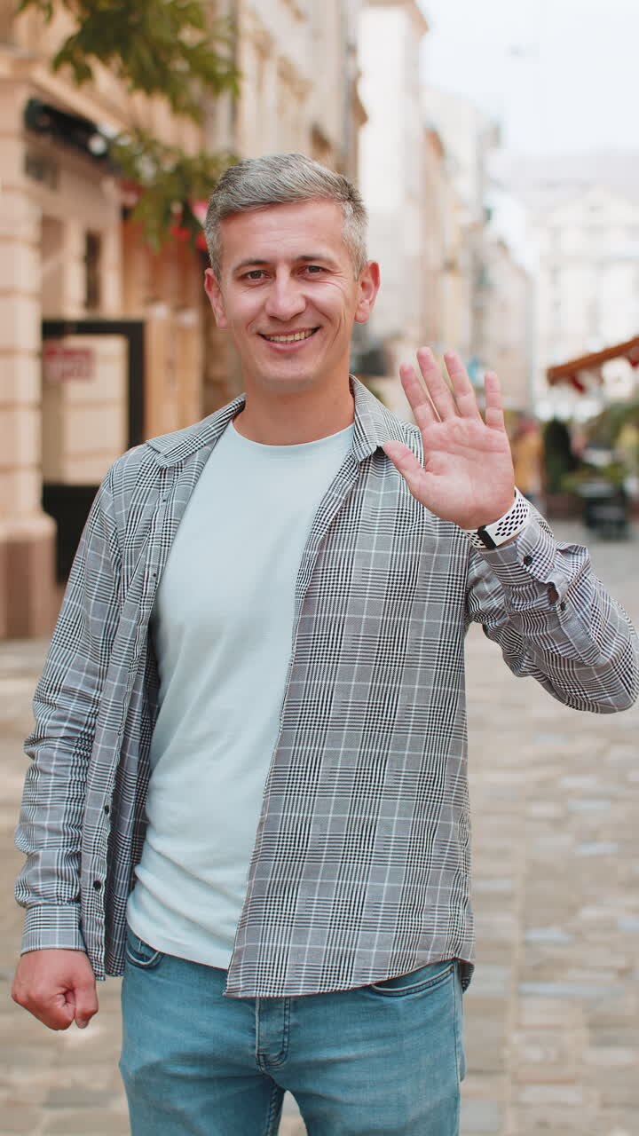 Uomo maturo allegro, turista che sorride alla telecamera, agita le mani, fa un gesto, saluta, in piedi per strada.