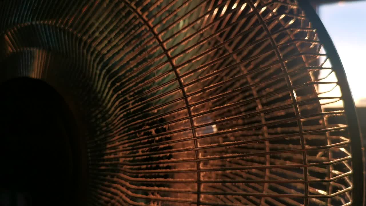 Dusty indoor fan during sunset. Low sun breaking through grid and propellers of ventilator causing lens flares. Slow upwards camera movement revealing sun shining through window.