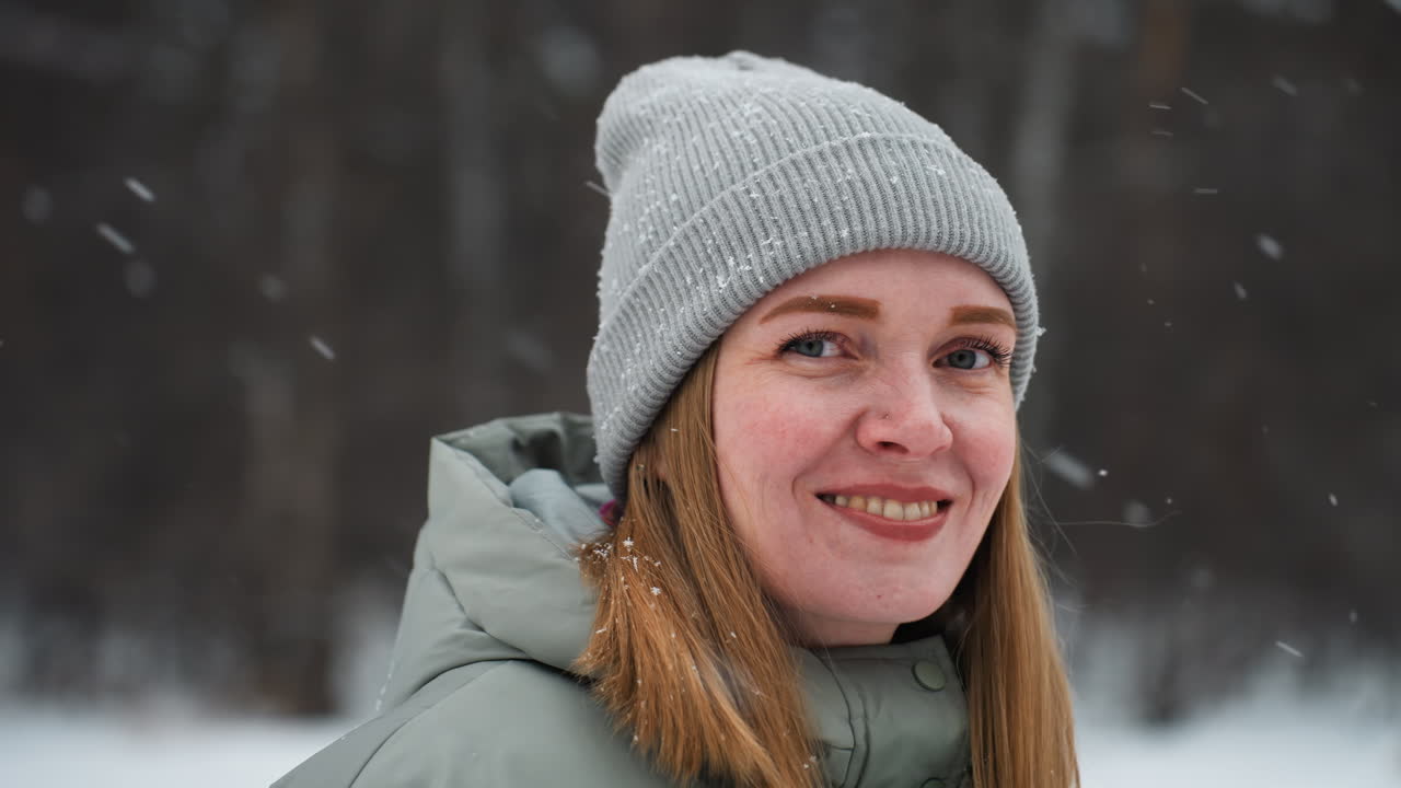Smiling woman wearing gray knit beanie and light winter coat standing outdoors during snowfall in snowy forest, with snowflakes visible on clothing and hair, expressing warmth and joy in cold weather