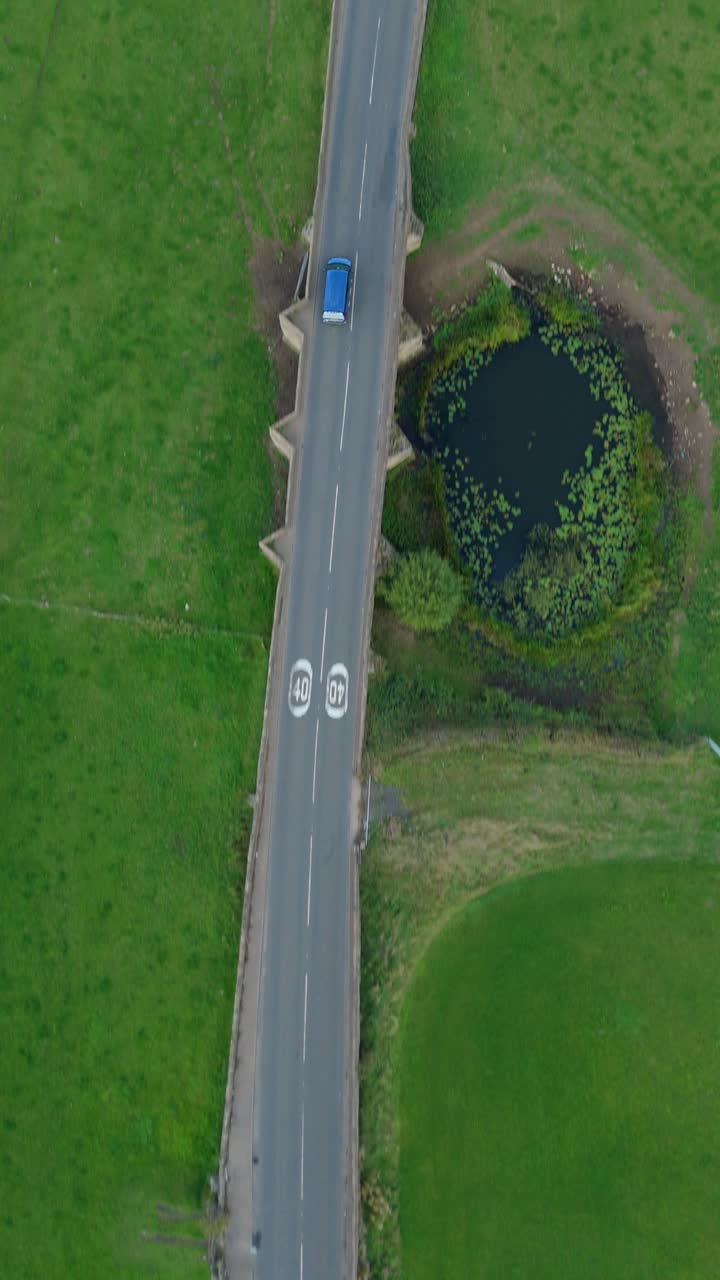 Overhead cinematic drone pass of bridge spanning river with reedbeds mudflats and vehicles moving through Derbyshire countryside
