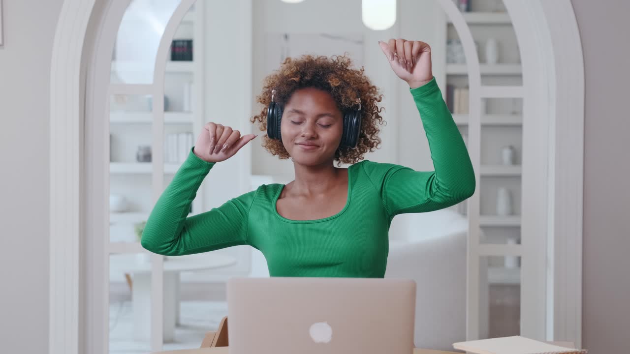 Giovane donna afroamericana con le cuffie balla seduta al tavolo con il laptop