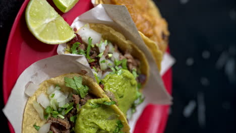 vertical slow motion of a red plastic plate with variety tacos at a traditional mexican restaurant with a lime sliced on the side and guacamole on top