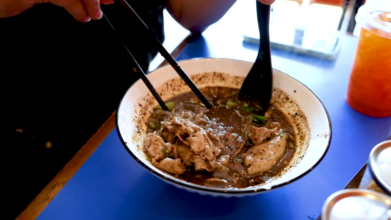 Lähikuva käsistä nostamassa nuudeleita syömäpuikoilla tummasta mausteisesta naudanlihakeitosta