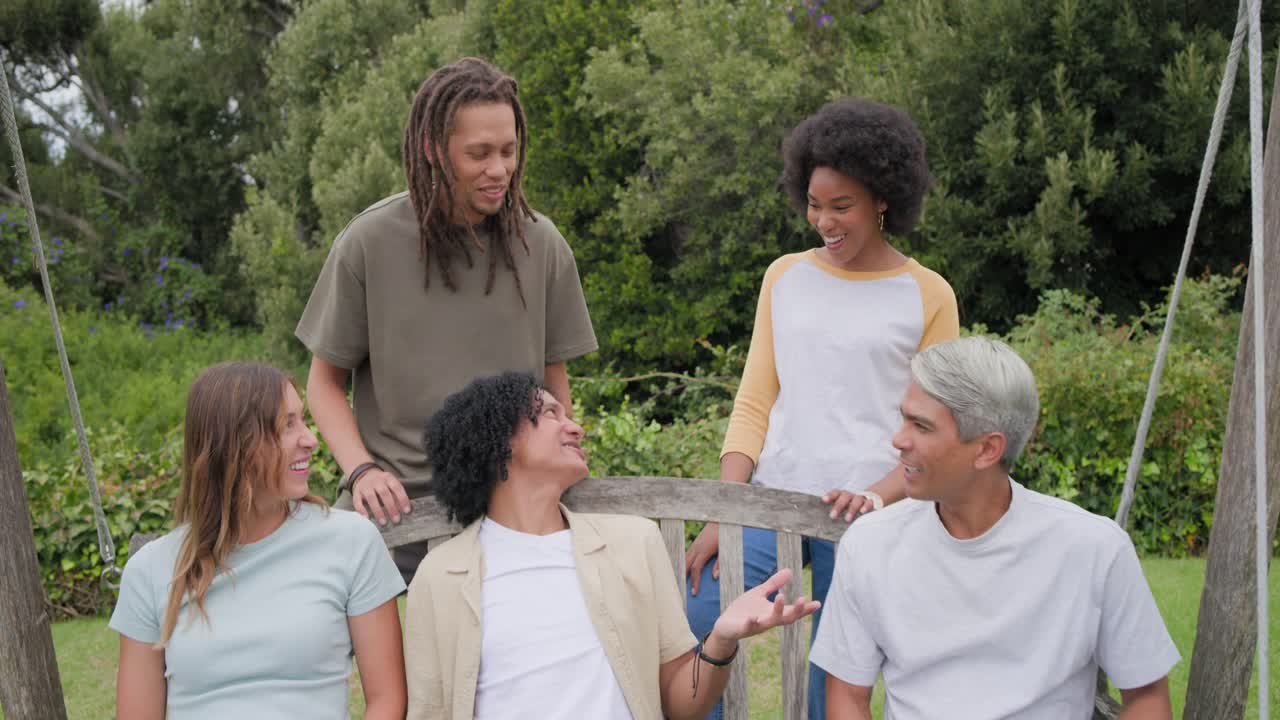Family sitting on wooden swing bench on grass, central man initiating conversation, group laughing
