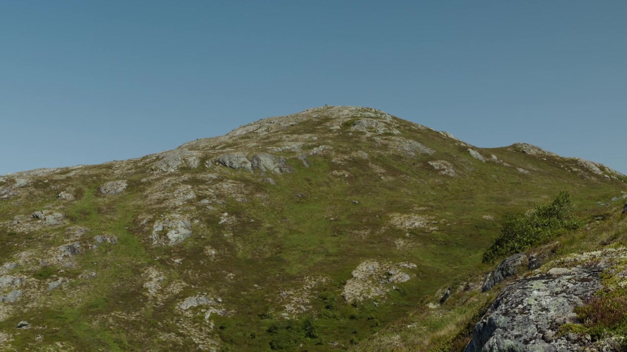 Una collina erbosa e rocciosa delle alture di Sunnmøre si erge sotto un cielo senza nuvole in una luce diurna nitida, suggerendo una remota natura selvaggia nordica