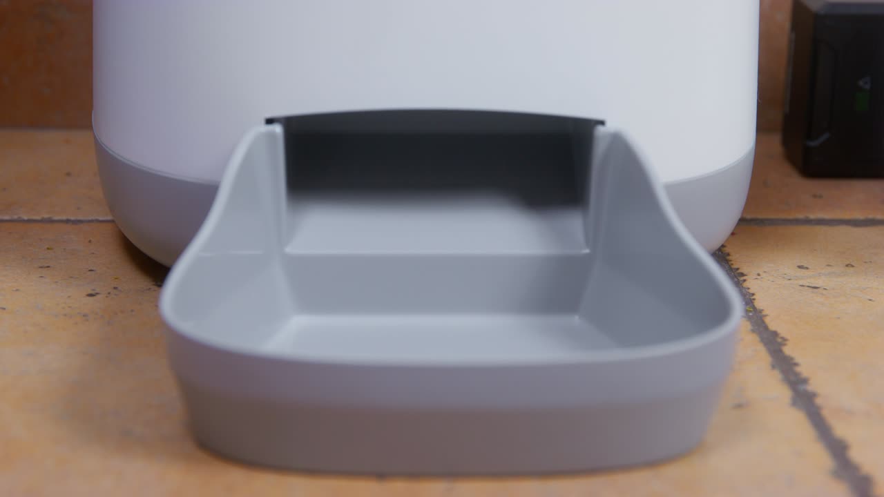 moderno alimentador automático de plástico blanco para mascotas de pie en el piso de azulejos naranja en la cocina contra la pared de yeso muy de cerca