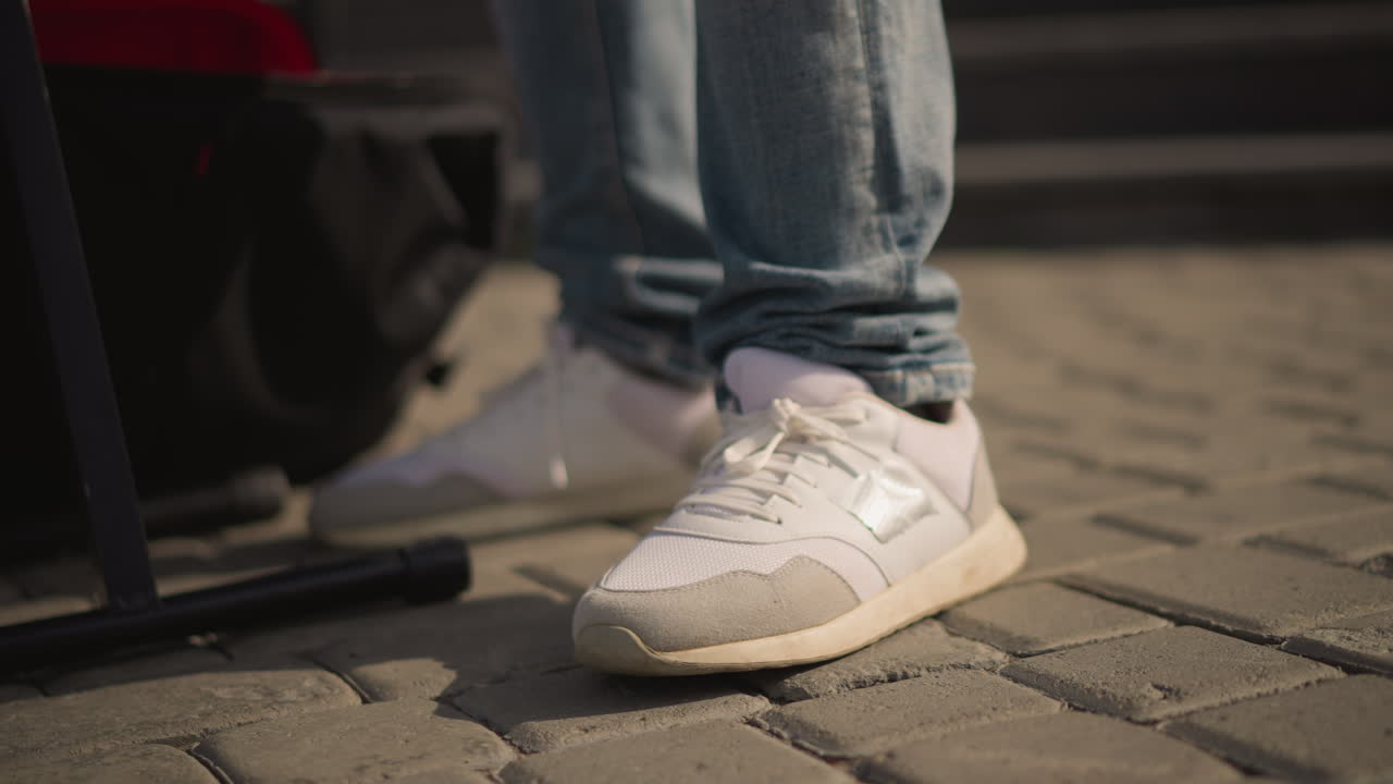 Hvide sneakers på brolagt gade, nærbillede af jeansklædte ben ved siden af keyboardstativ og rygsæk, diskret fodtappen og hælløft, varmt eftermiddagslys, intim bymusiker-stemning.
