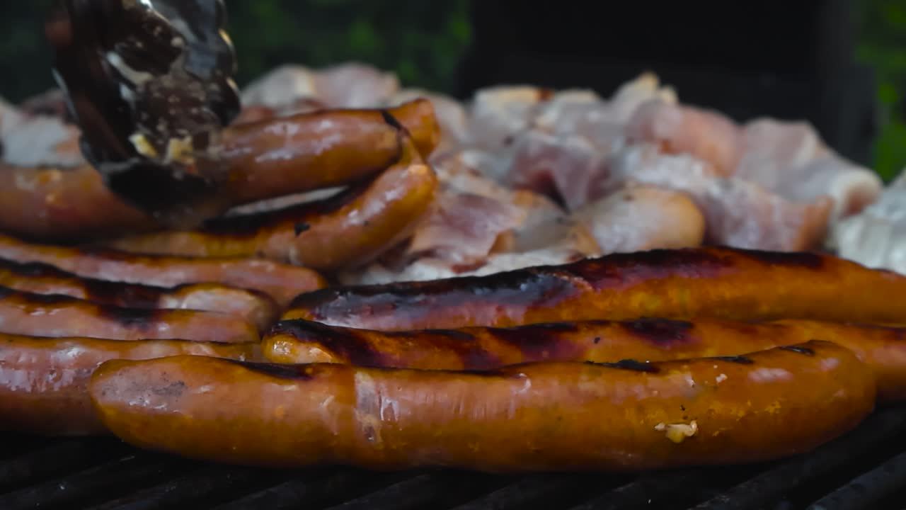 Close up of caramelized sausages and bacon sizzle over hot charcoal grill with rising steam during outdoor summer barbecue. Juicy meat with flame grilled stripes while chef turns the food with tongs