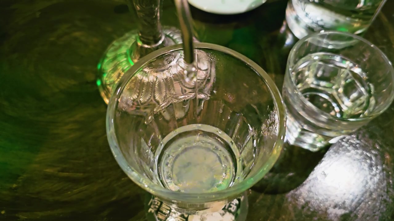 Primer plano cenital de la formación de louche de absenta en un vaso mientras gotea agua fría de un caño de fuente, con líquido pálido y turbio, una mesa de bar reflectante e iluminación ambiental verde de salón.