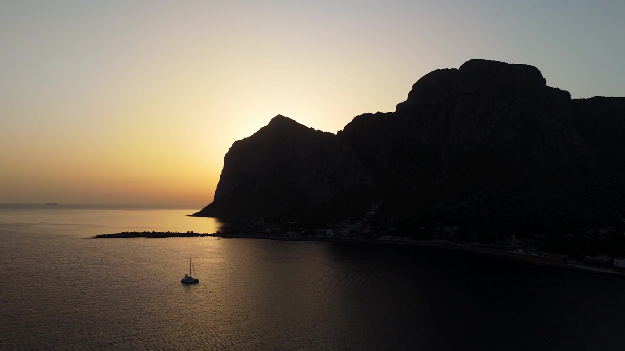 Luftfoto fra drone af silhuetten af den klippefyldte halvø Capo Gallo i Palermo, Italien, der tårner sig op over det rolige Middelhav under en strålende gylden solopgang