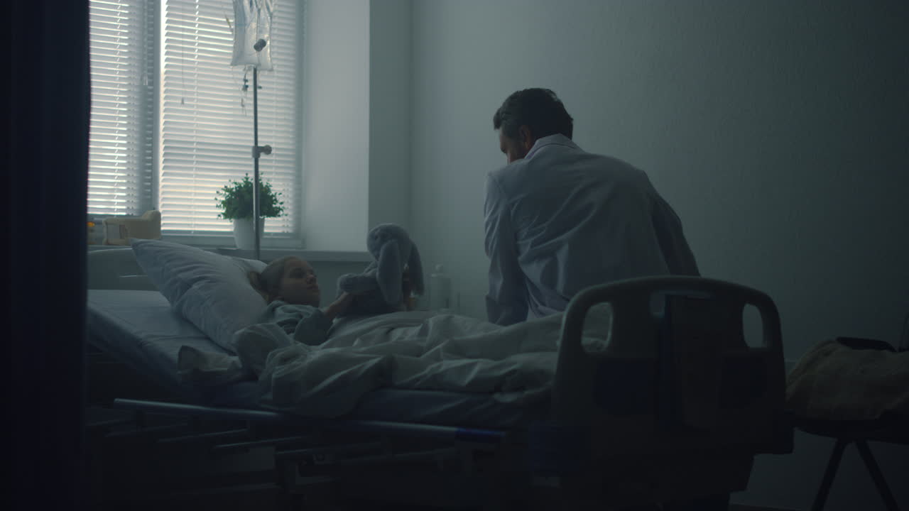 クリニックの病棟で小さな患者を訪問する医師 ⁇ 病気の女の子の抱きしめ ⁇ ベッドでふわふわの玩具 ⁇