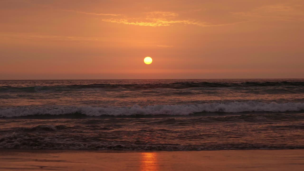 Sunset seen from the shore. Waves crash onto the sand as the sky turns orange and the sun sets. The water on the sand reflects the sun's rays.