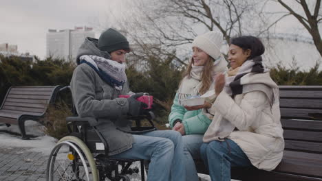 happy disabled man in wheelchair opening a gift box while celebrating his birthday with two friends at urban park in winter