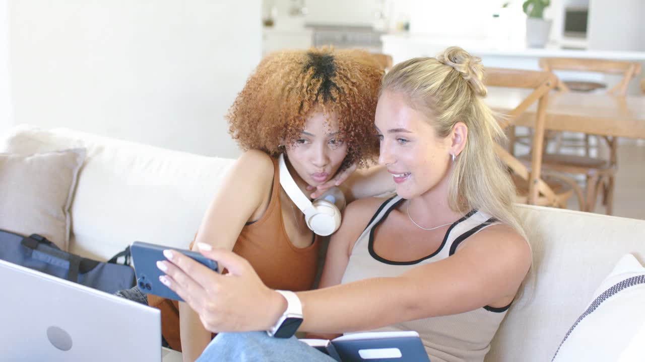 Posing two women in beige tank and brown sleeveless, scrolling smartphone on sofa at home, laptop