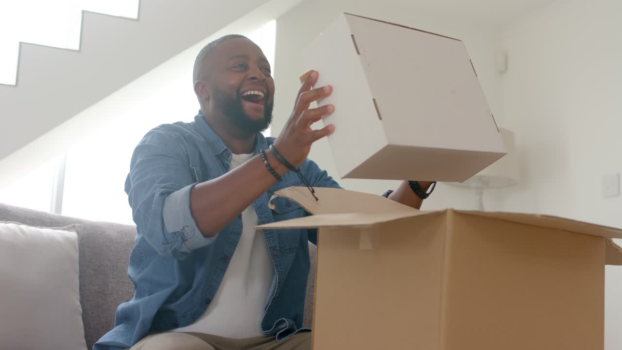 African American man sitting on sofa opening big package pulling white box checking items laughing