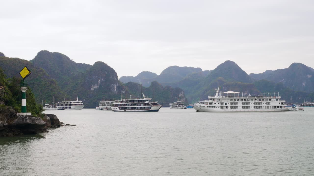 Majestic limestone karsts and calm waters of Ha Long Bay create a stunning backdrop for multiple white luxury cruise ships anchored in the tranquil lagoon during an overcast day