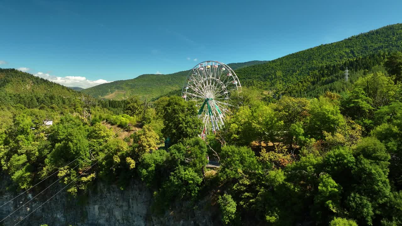 A ferris wheel stands tall above a deep forest canyon, surrounded by rich vegetation and distant mountain silhouettes under clear skies