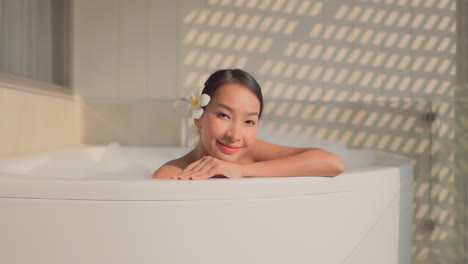Close-up-of-a-young-Asian-woman-leans-on-the-edge-of-a-resort-style-bathtub-looking-out-from-her-luxurious-bubble-bath