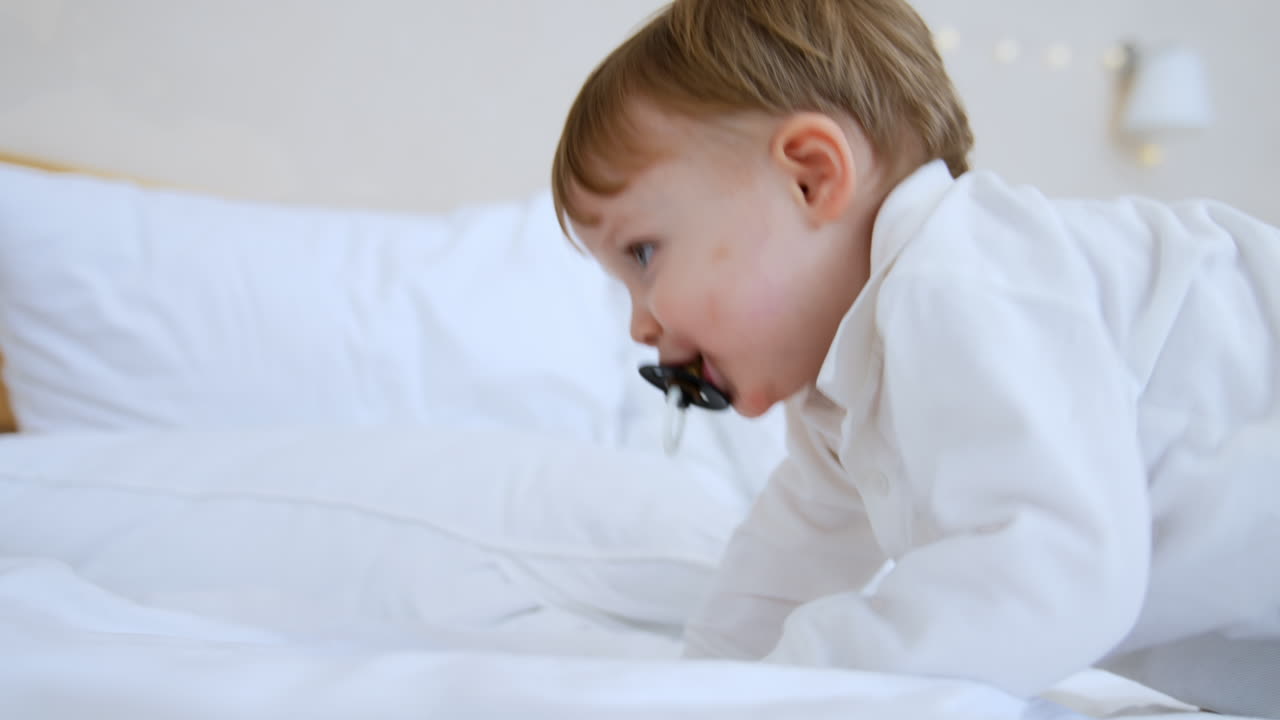 Cute grey-eyed blond Caucasian toddler with a pacifier lying on the bed. Baby drops a pacifier, gets it back and climbs off the bed.