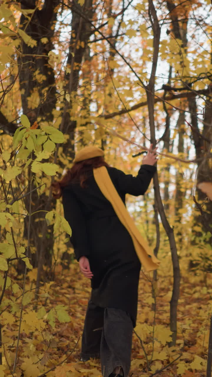 vista trasera de una mujer con una boina amarilla y abrigo negro, caminando a través de un denso bosque de otoño, rodeado de hojas amarillas vibrantes