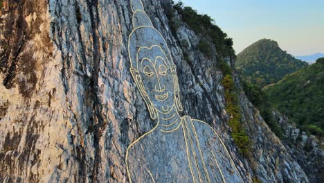 Vista-Aérea-De-4k:-Imagen-De-Buda,-Sentado-Con-Las-Piernas-Cruzadas,-Grabado-Con-Oro-En-La-Cara-Norte-De-Una-Colina-De-Piedra-Caliza-En-Khao-Chi-Chan,-Tailandia