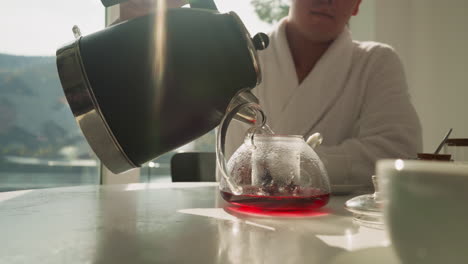 man pours water from kettle into teapot. fruit tea brewed by gradually filling with water with help of young man. dining table with hot beverage