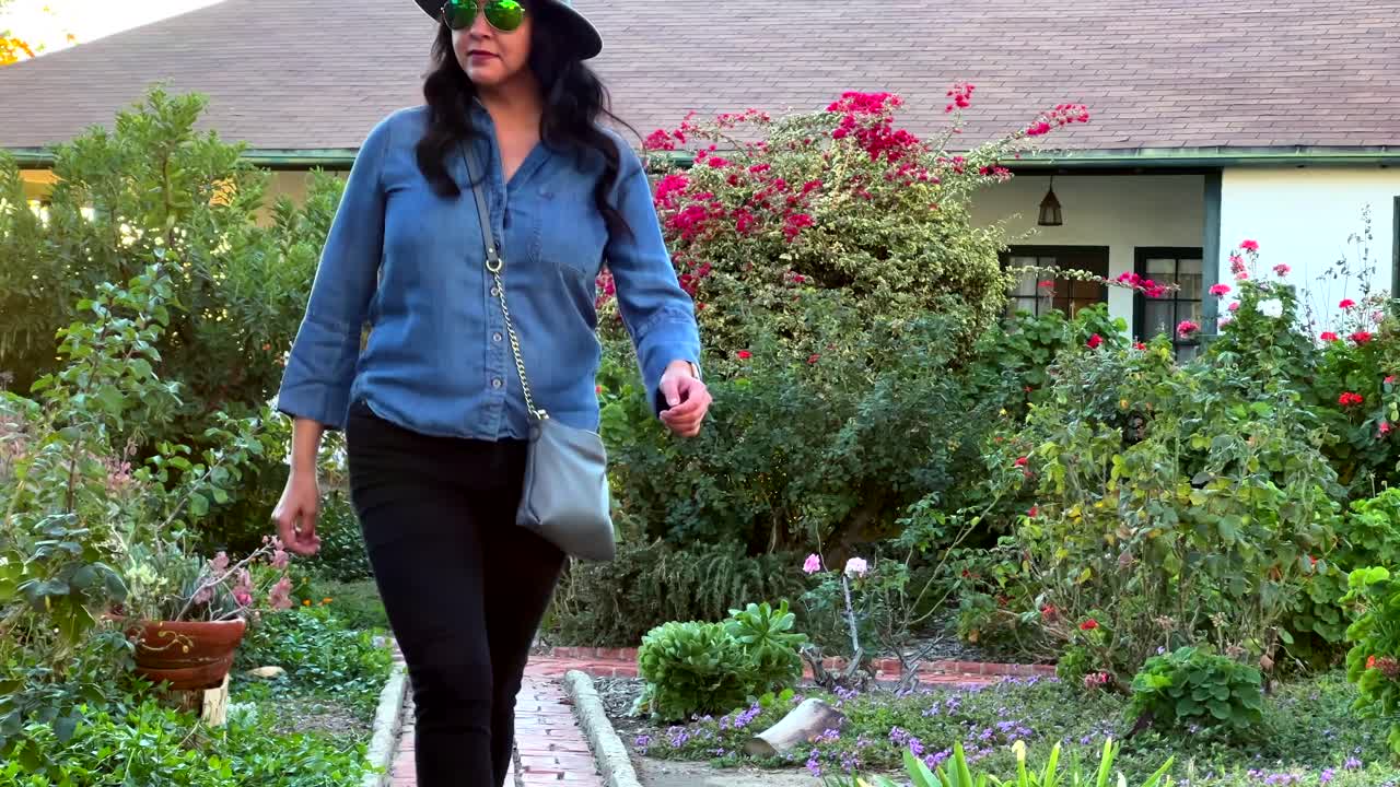 mujer de moda caminando por un colorido jardín con gafas, sombrero y bolso en un día soleado