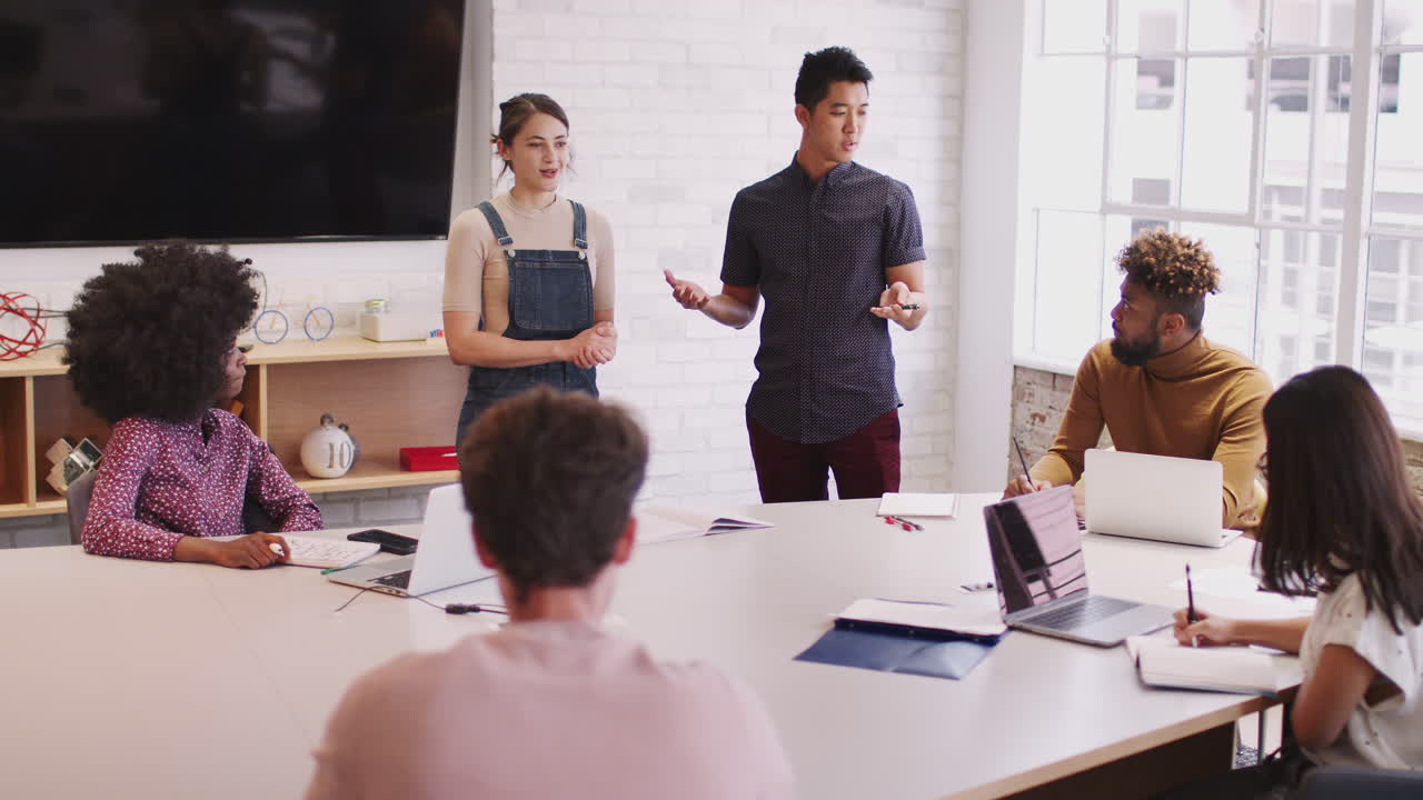 Two millennial colleagues stand presenting to creative team in a meeting room