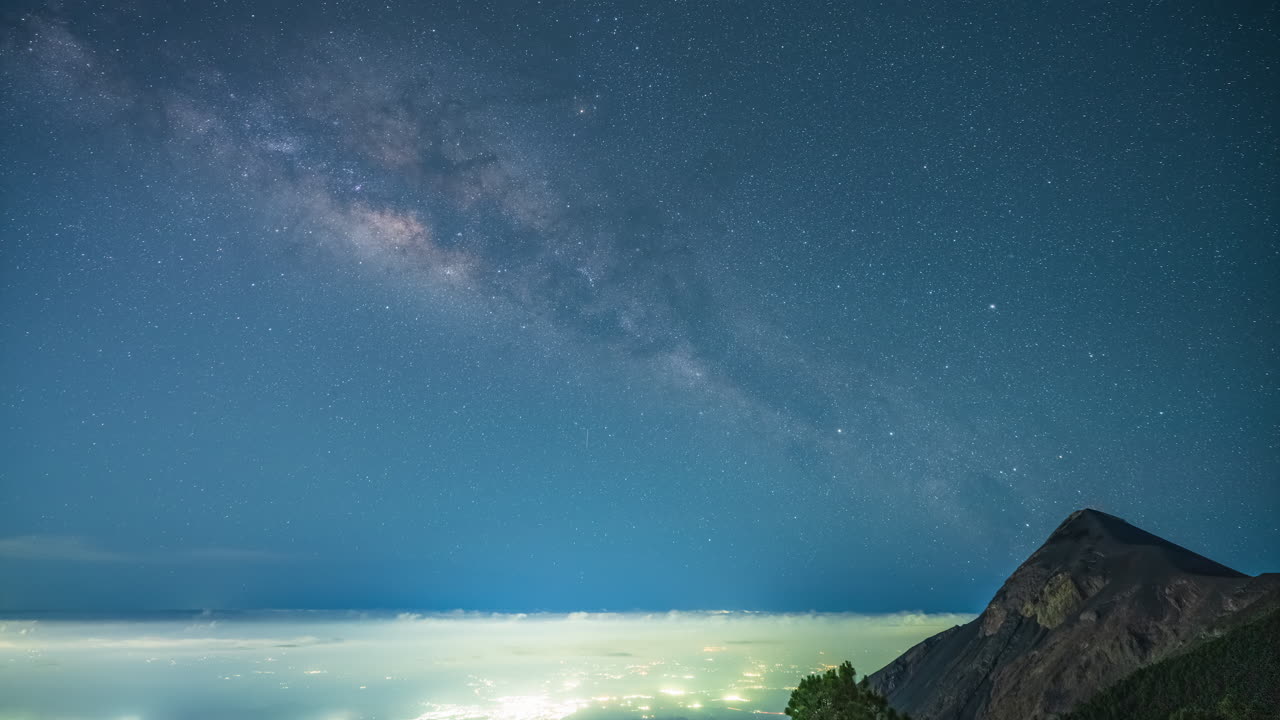 Time lapse of the Milky Way stretching above the active Fuego Volcano in Guatemala, with glowing city lights below and a vivid, starry night sky