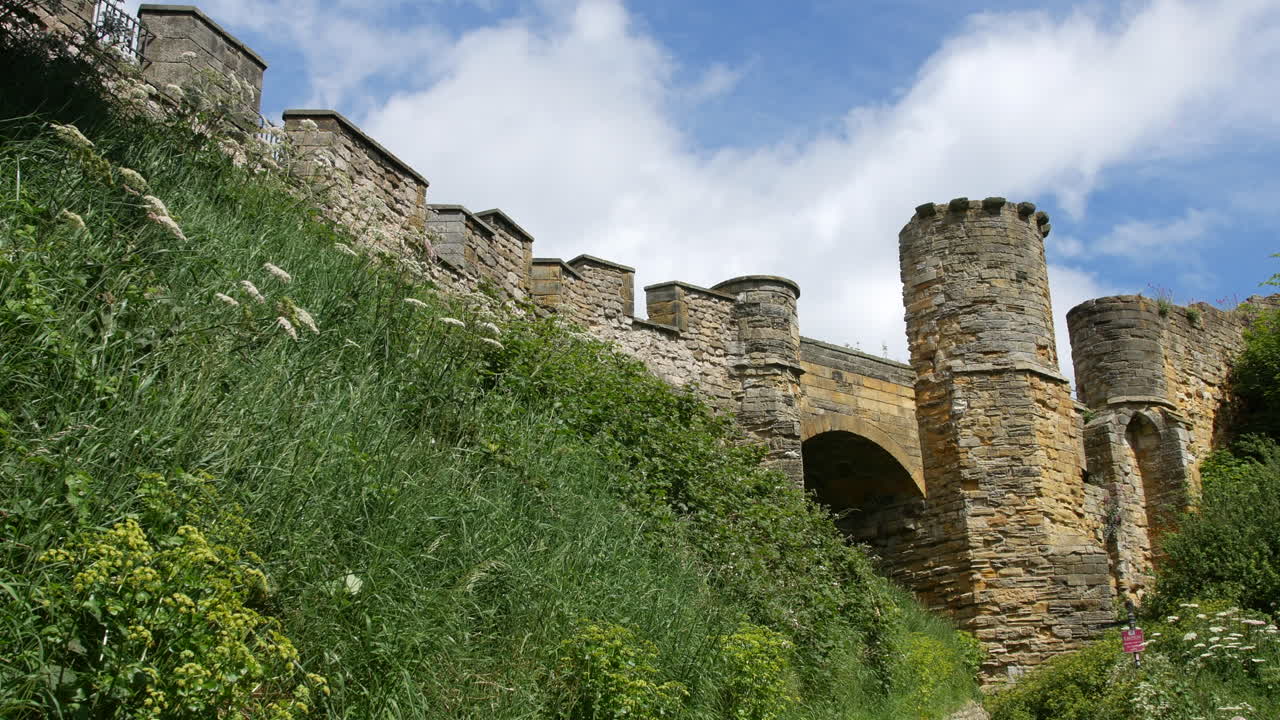Castle walls and towers rise above a green slope in Scarborough, North Yorkshire, England, with sunlight illuminating the stone structure between trees