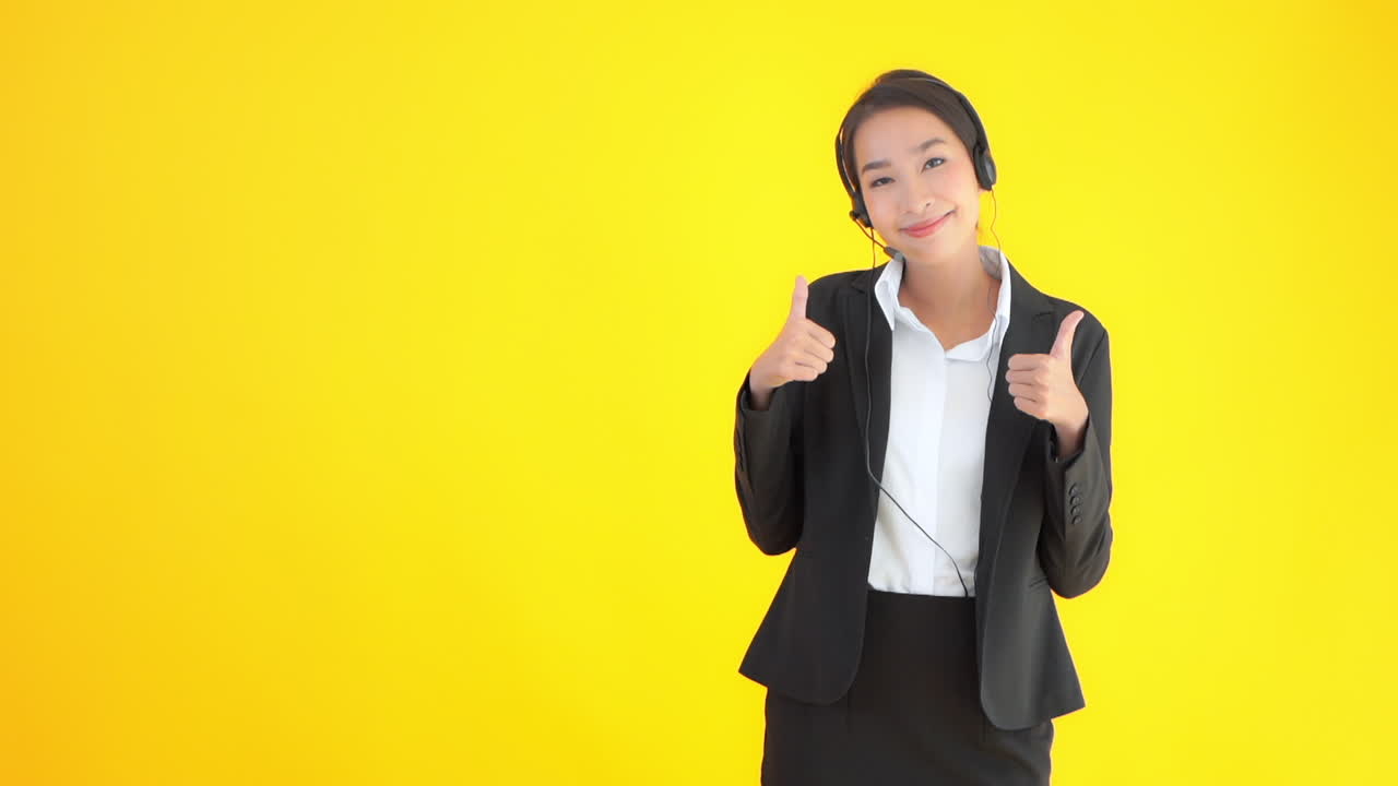 An attractive young Asian woman wearing a business suit and a headset with a microphone gives the thumbs up for success. Copy space