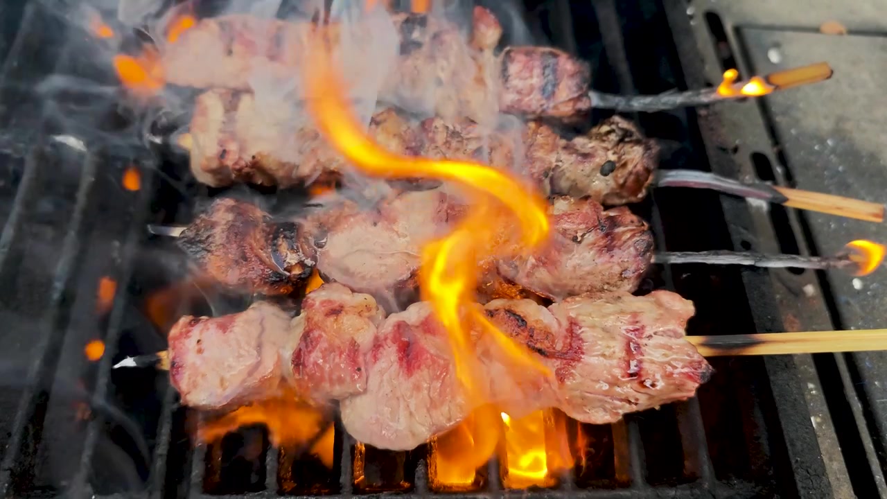 Video orizzontale in slow motion 4K di spiedini di manzo in preparazione su una griglia per barbecue, con fiamme, fumo e carne che sfrigola, che cattura la cucina all'aperto, la grigliata e la preparazione di cibo delizioso