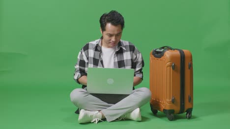 cuerpo lleno de viajero masculino asiático con equipaje usando una computadora portátil mientras está sentado en el estudio de fondo de pantalla verde