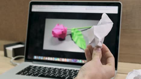 person holding crumpled paper in front of a laptop screen displaying a pink origami rose.