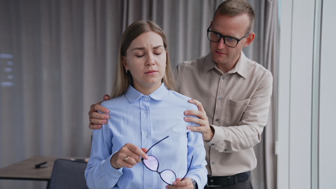 Man is holding a lady by her shoulders from behind. Lady feeling bad and rubs her nose bridge. Male colleague talks and comforts lady.