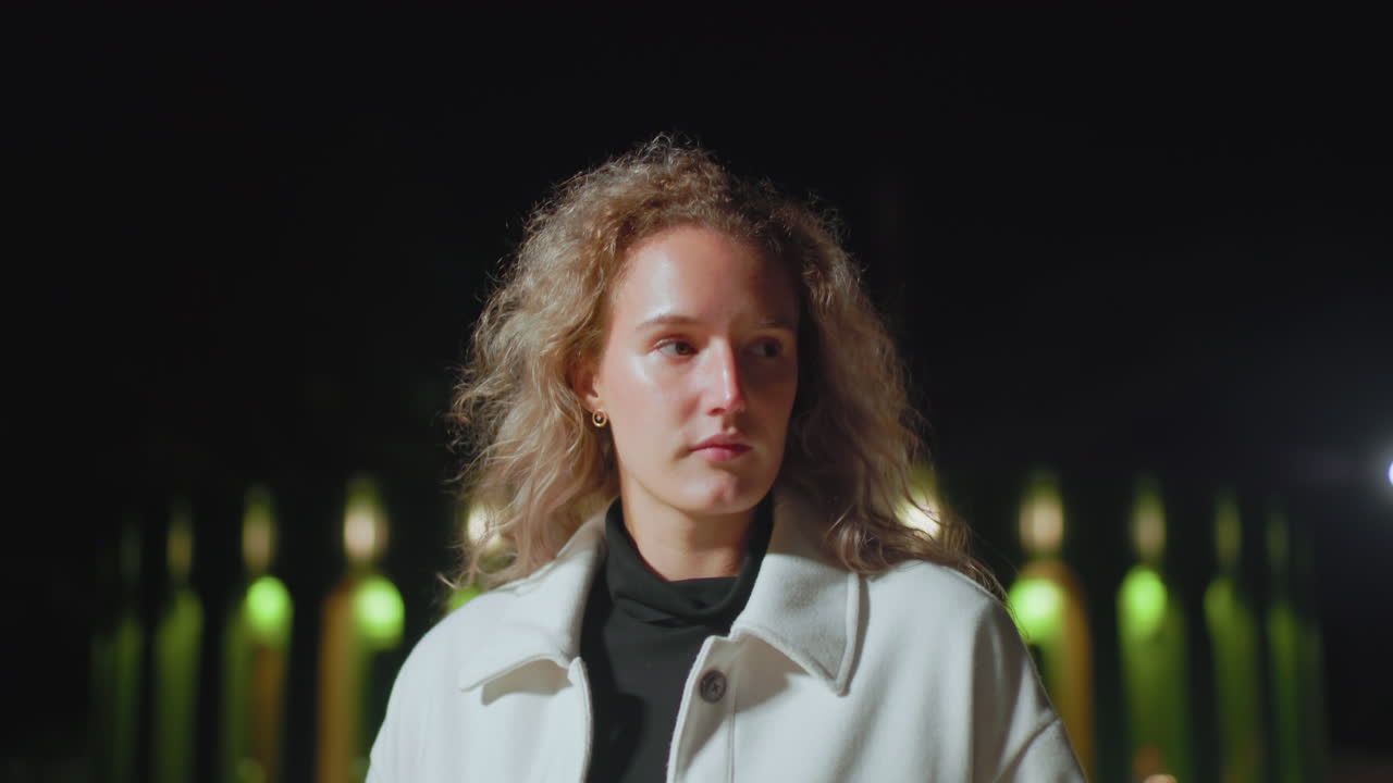 Portrait view of woman in white coat walking calmly outdoors at night with eyes closed, illuminated by soft glowing lights in background, creating serene mood in quiet nighttime environment