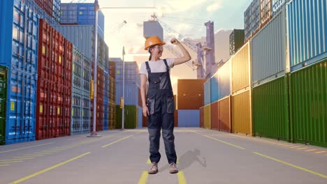 cuerpo lleno de mujer asiática trabajadora con gafas de protección y casco de seguridad sonriendo y flexionando su bíceps mientras estaba de pie en el depósito de contenedores