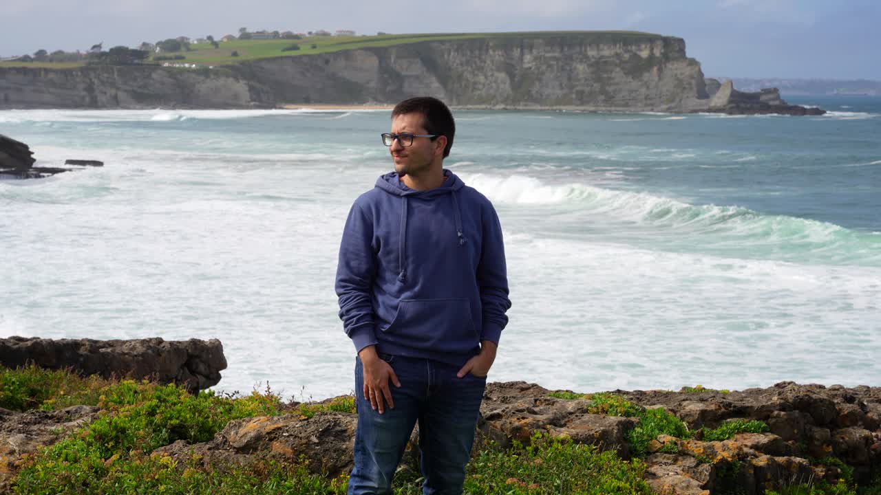 Young man in a hoodie stands by the cliffs of Galizano with waves crashing against the rocky shore