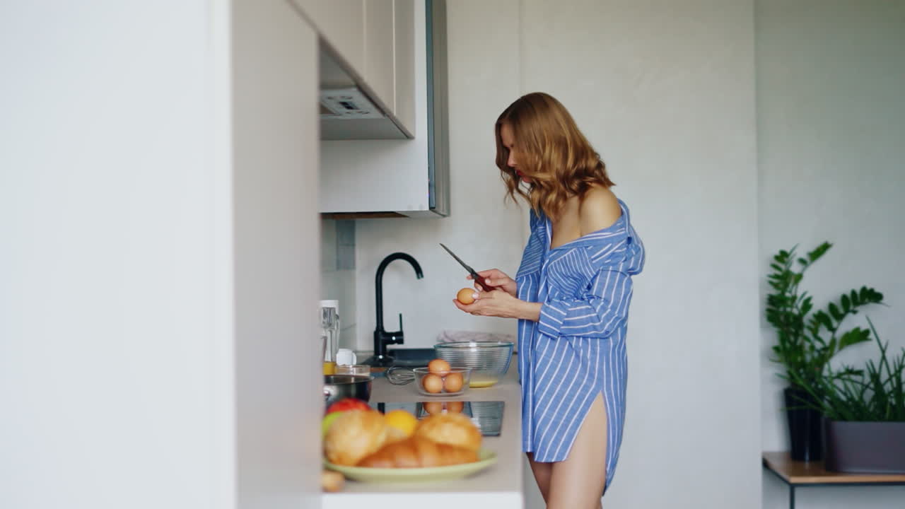 chica sexy en camisa rompiendo huevos en un cuenco de vidrio en la cocina