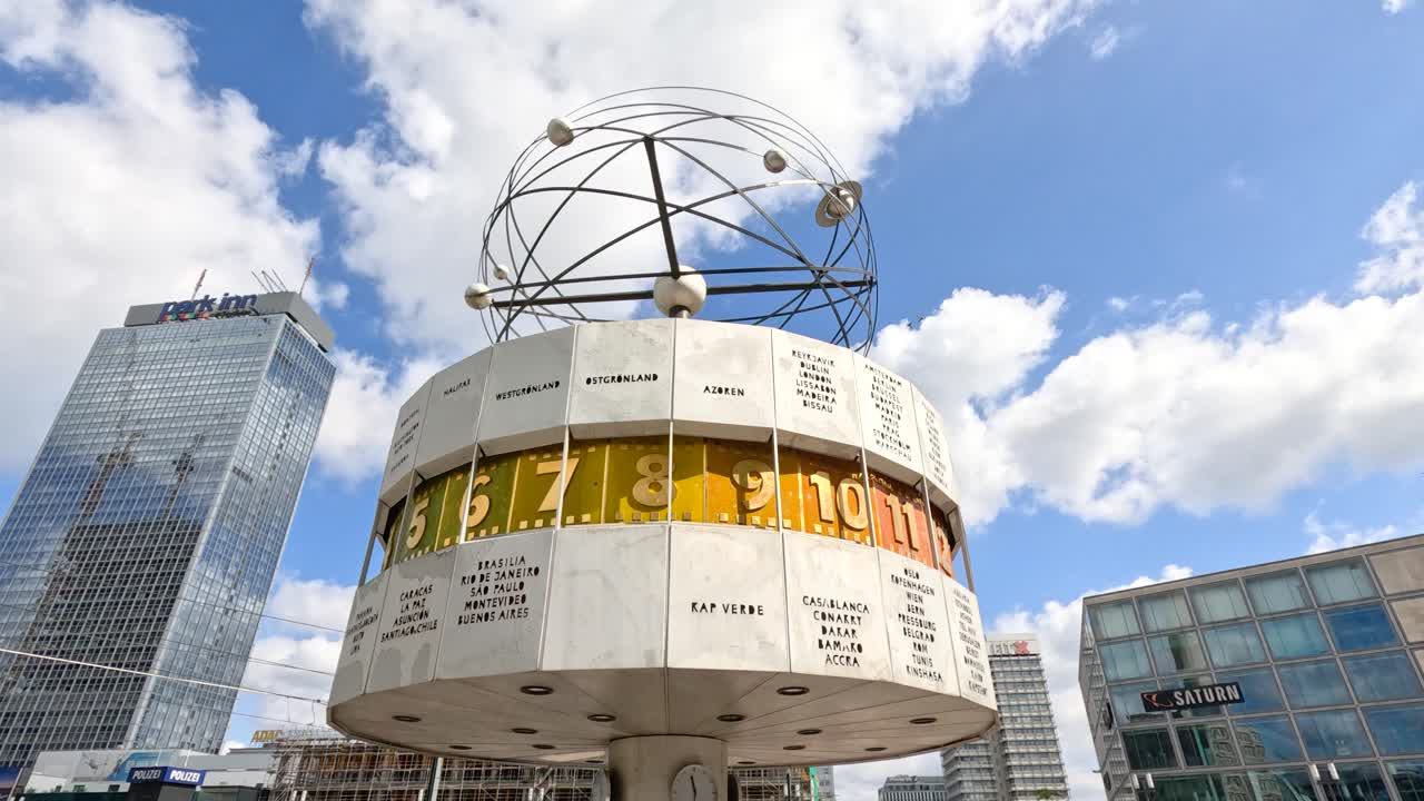Low-angle video of rotating world clock at city plaza with modern buildings and blue sky