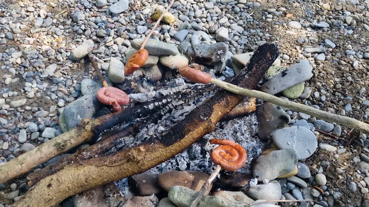 İsviçre’de Grillschnecken olarak bilinen dört adet spiral sosis, rustik bir açık hava pişirme deneyimi sunarak parıldayan bir kamp ateşinde tahta çubuklarda pişiyor.