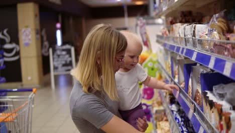 Young-mother-in-glasses-holding-her-child-in-her-arms-while-choosing-cookies-on-the-shelves-in-the-supermarket.-Thoughtful-mom