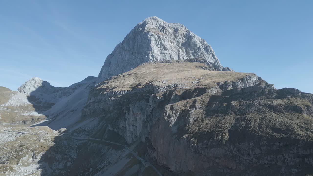 Drone view of Mangart Mountains, Slovenia: jagged peaks, rugged cliffs, lush green valleys, winding alpine roads, dramatic landscape, clear skies, Julian Alps, untouched natural beauty