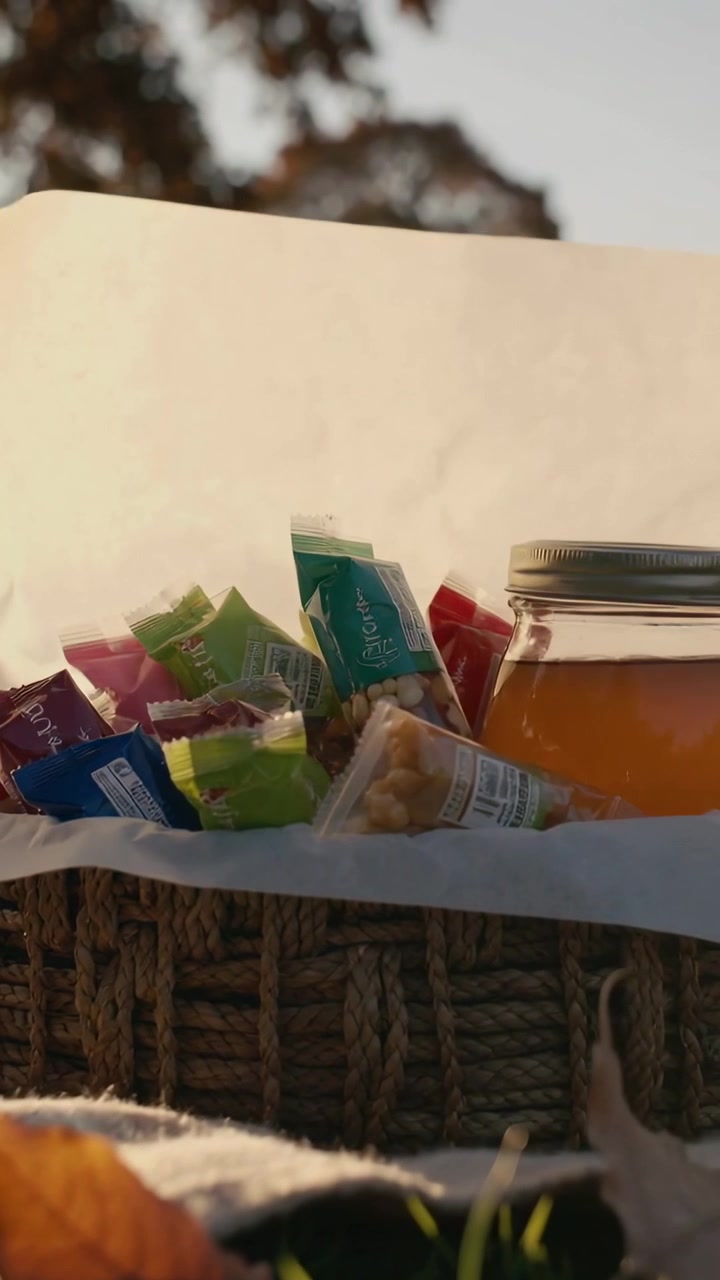 Vertical video: Lifting off-camera hand revealing wicker picnic basket at park to show snacks, jar