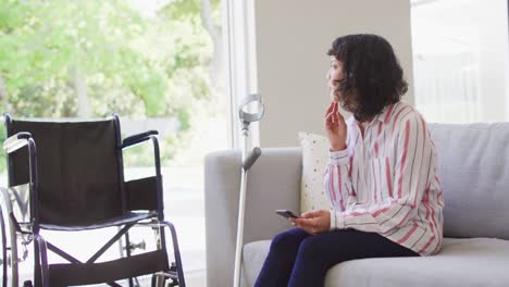 Thoughtful-biracial-disabled-woman-using-smartphone-in-living-room,-crutch-and-wheelchair-by-couch