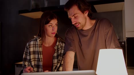 una pareja feliz un tipo moreno en una camiseta gris y su novia en una camisa a cuadros están viendo videos divertidos y fotos de su vida en la cocina en un apartamento moderno por la noche usando una computadora portátil