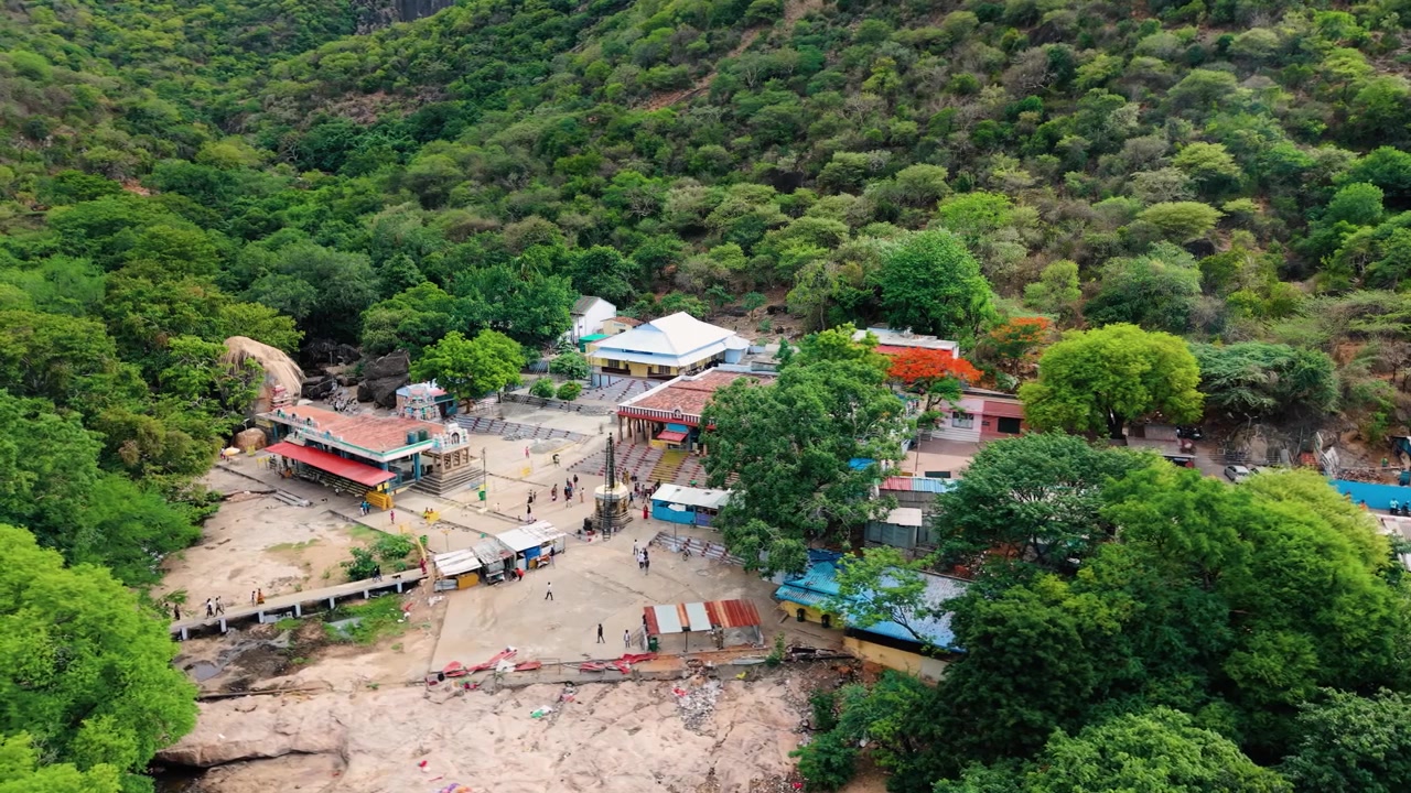 Pemandangan Drone Udara Sinematik Kompleks Kuil Kuno Thirumoorthy Hills yang Terletak di Kaki Bukit Western Ghats yang Berhutan, Umat Berjalan di Halaman, Lereng Gunung Hijau Lebat Mengelilingi Lokasi