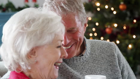 Nahaufnahme-Eines-älteren-Paares,-Das-Zu-Hause-Mit-Einem-Heißen-Getränk-Auf-Dem-Sofa-Sitzt-Und-Sich-Mit-Dem-Weihnachtsbaum-Im-Hintergrund-Unterhält